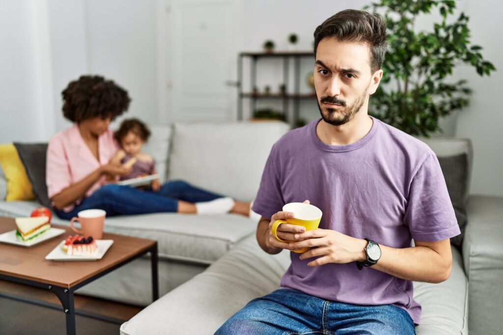 Man holding a mug looking concerned or skeptical with his family on a sofa behind him.