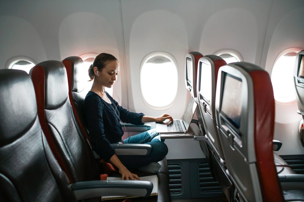 Woman on an airplane with the row to herself as she works on her laptop and looks at a notebook in the seat next to her.
