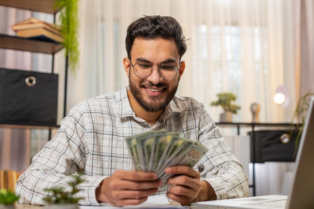 Happy man counting cash and using laptop in home office.