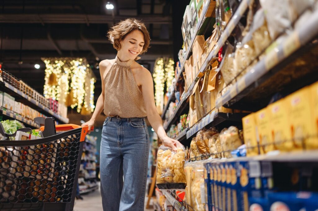 3 Regional Grocery Stores That Shoppers Love Even More Than Costco in 2026 Smiling young woman picking up a package of pasta in a grocery store, pushing a cart.