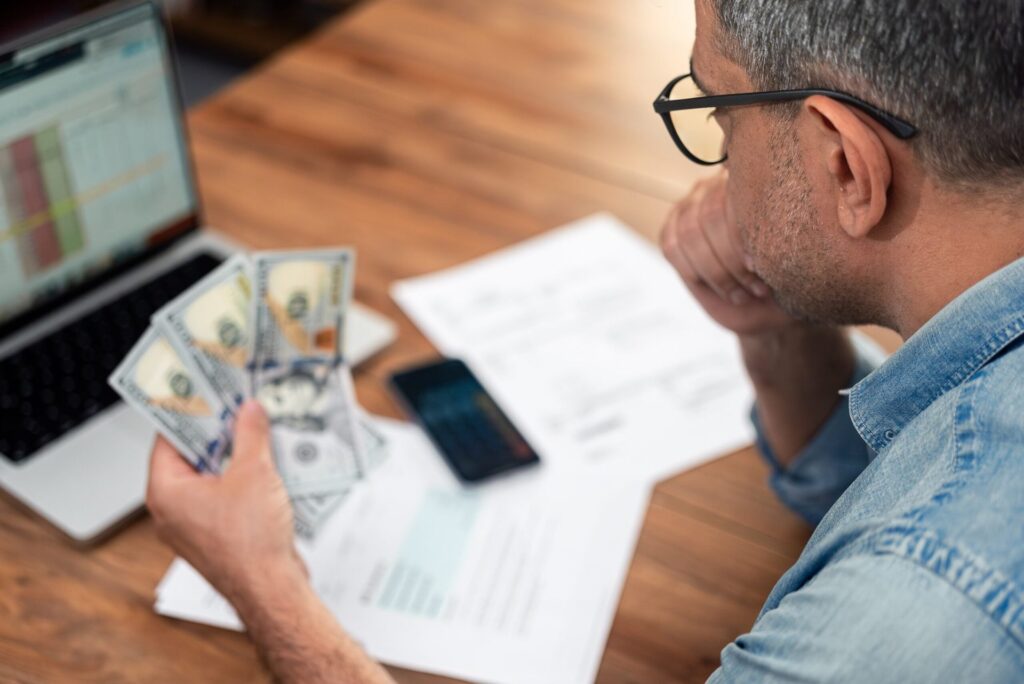 Older man looks over his budget on a laptop while counting cash.