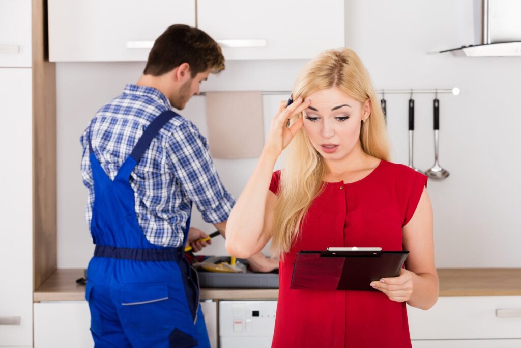Plumbers Beg You: Stop Flushing These 5 ‘Safe’ Items or Face a $4,000 Bill Stressed woman signing a document or bill for plumbing while a plumber packs up his tools in the kitchen